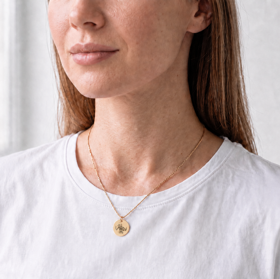 A woman wearing a delicate gold necklace with a small round pendant, styled with a white crew-neck t-shirt in a bright, softly lit indoor setting.