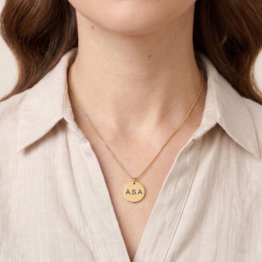 A close-up of a woman wearing a delicate gold chain necklace with a round gold disc pendant engraved with the initials 'ASA'. She is wearing a light beige linen button-down shirt against a soft, warm neutral background.