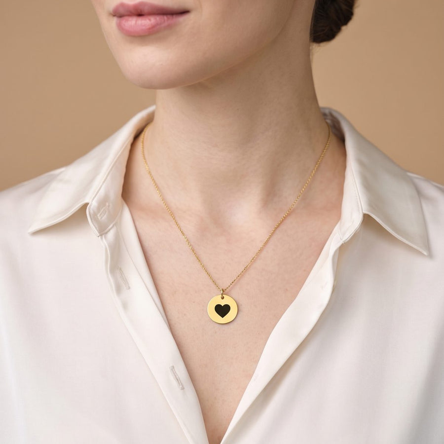A close-up of a woman wearing a delicate gold chain necklace with a circular gold pendant featuring a black heart in the center, paired with a white collared blouse against a warm beige background.