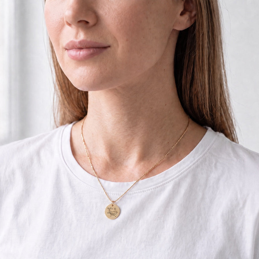 A woman wearing a delicate gold chain necklace with a small round gold coin pendant, styled with a white crewneck t-shirt.