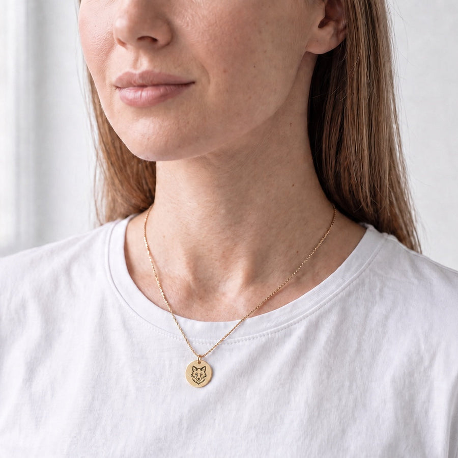 A woman wearing a delicate gold chain necklace with a small round engraved pendant, paired with a white crew-neck t-shirt. The background is a soft, bright, out-of-focus indoor setting.